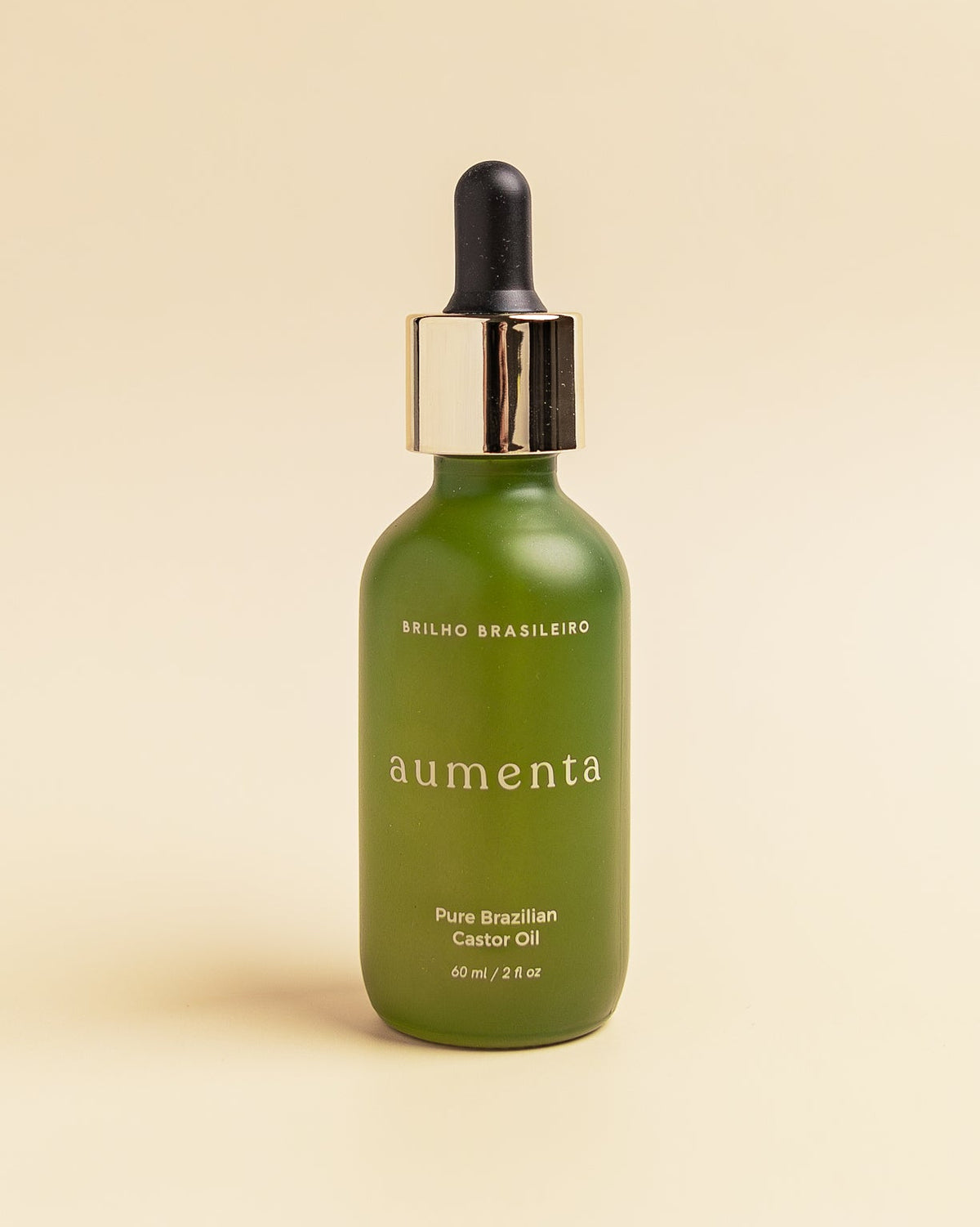 Aumenta Brilho Brasileiro castor oil in a green 60 ml glass bottle with a gold dropper cap, on a cream background